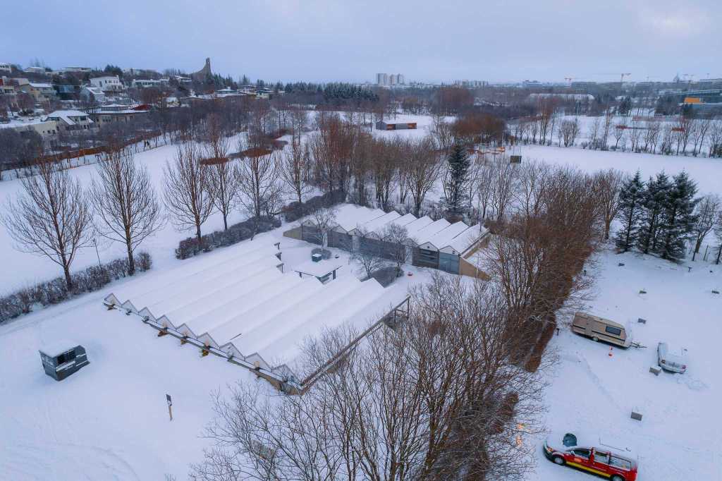 Snow-covered landscape with rows of greenhouses encircled by bare trees. Nearby, a red vehicle and a parked van rest under the overcast sky.