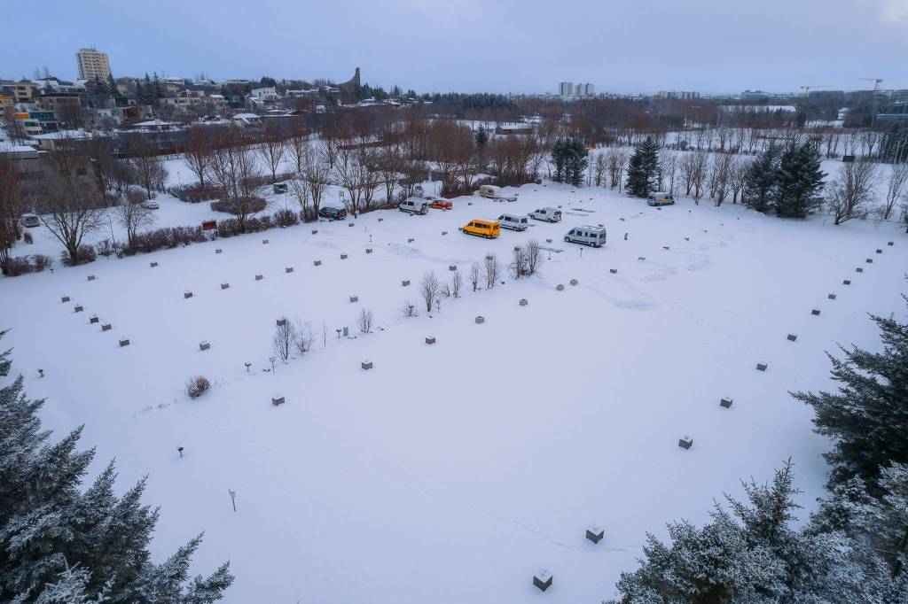 Snow-covered park with scattered concrete blocks and leafless trees, featuring brightly colored vehicles, under a gray, overcast sky.