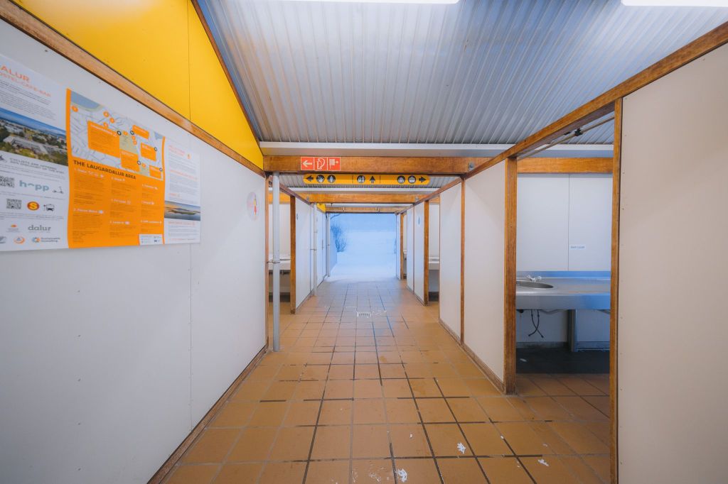 Hallway with yellow and white walls, tiled floor, and exits at each end. Posters adorn the left wall, and a stainless steel counter is on the right.