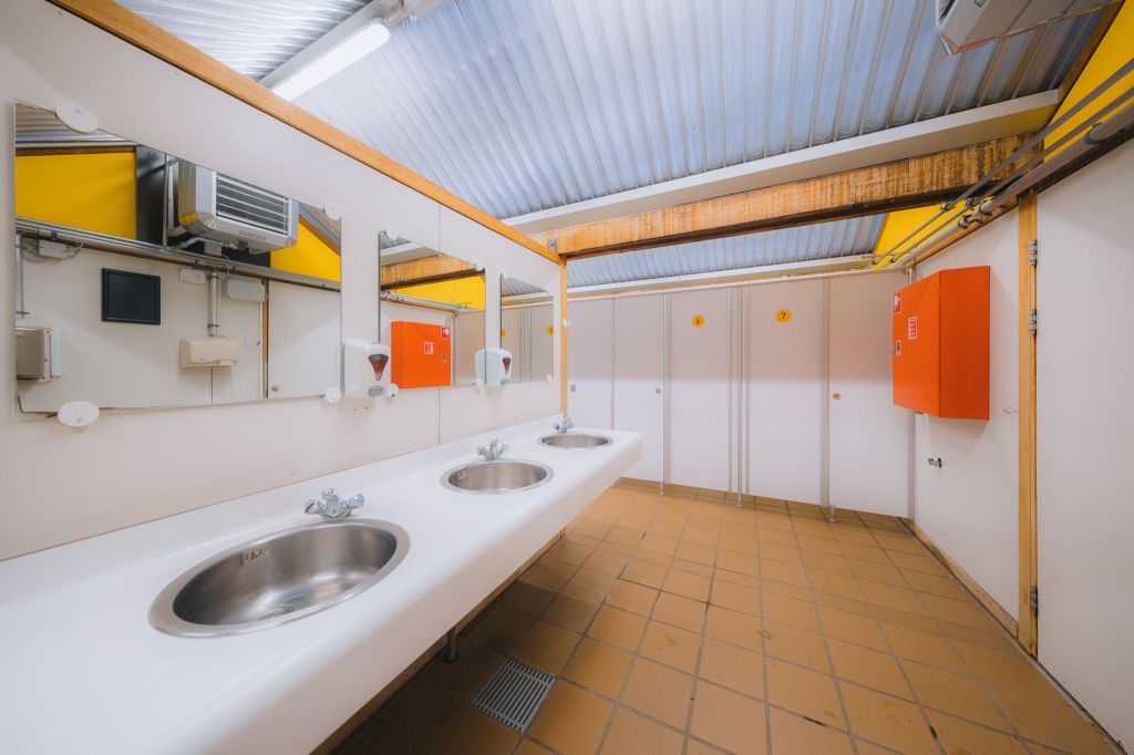 Public restroom with a clean, modern design. Features three metal sinks, mirrors above, white walls, and orange accents.