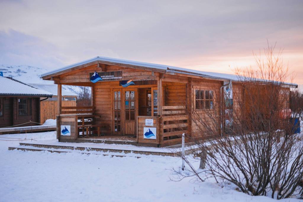 A small wooden cabin sits on a snowy landscape under a cloudy sky at sunset. The warm light from the cabin creates a cozy contrast against the cold surroundings.