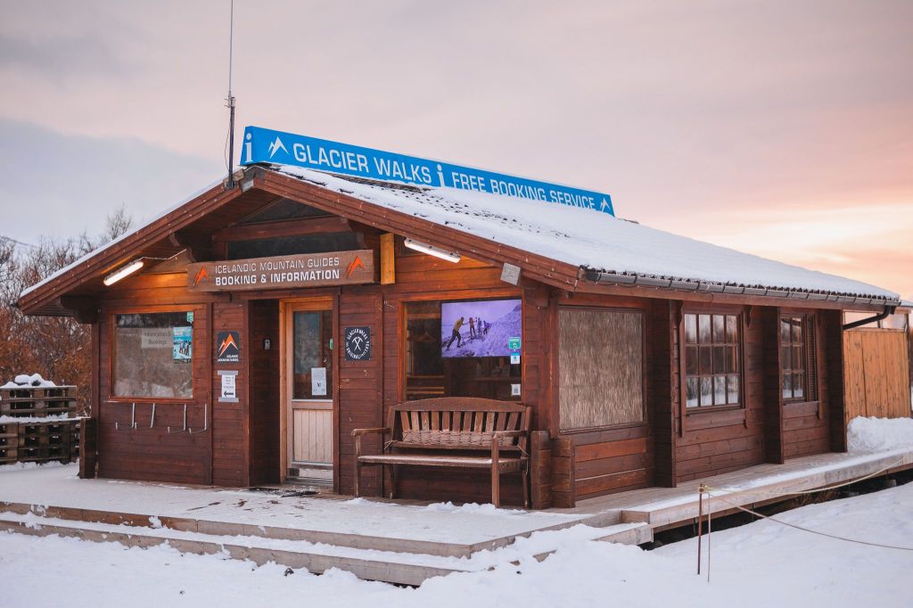 A cozy wooden cabin in snowy surroundings offers glacier walk bookings. A blue sign reads “GLACIER WALKS”.