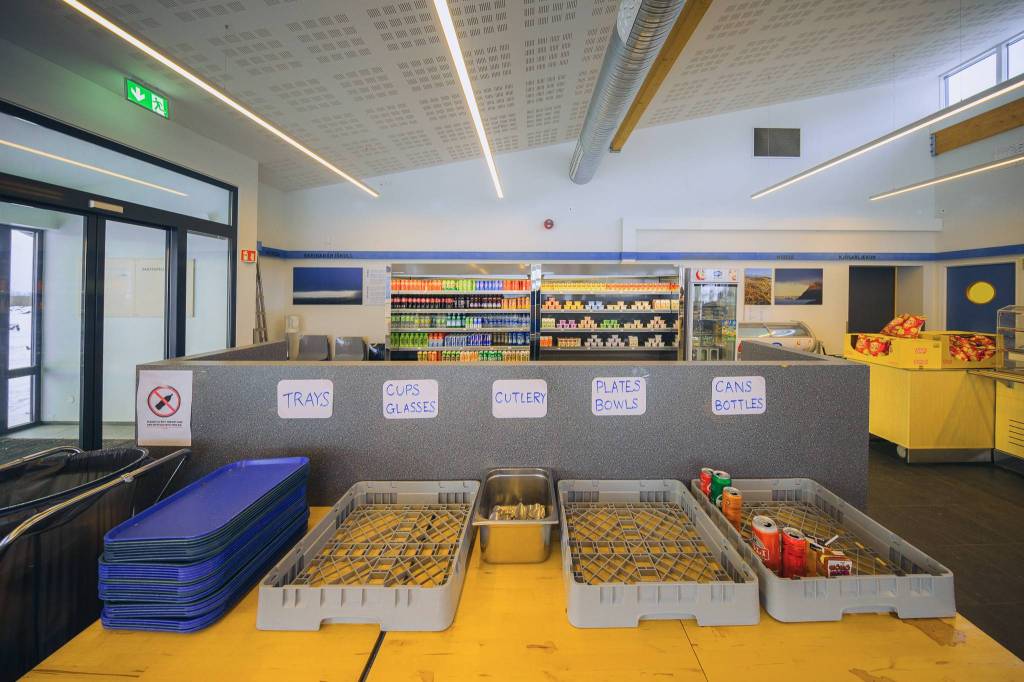 Cafeteria interior with recycling station labeled for trays, cups, cutlery, plates, and cans. Shelves stocked with drinks and snacks in the background.