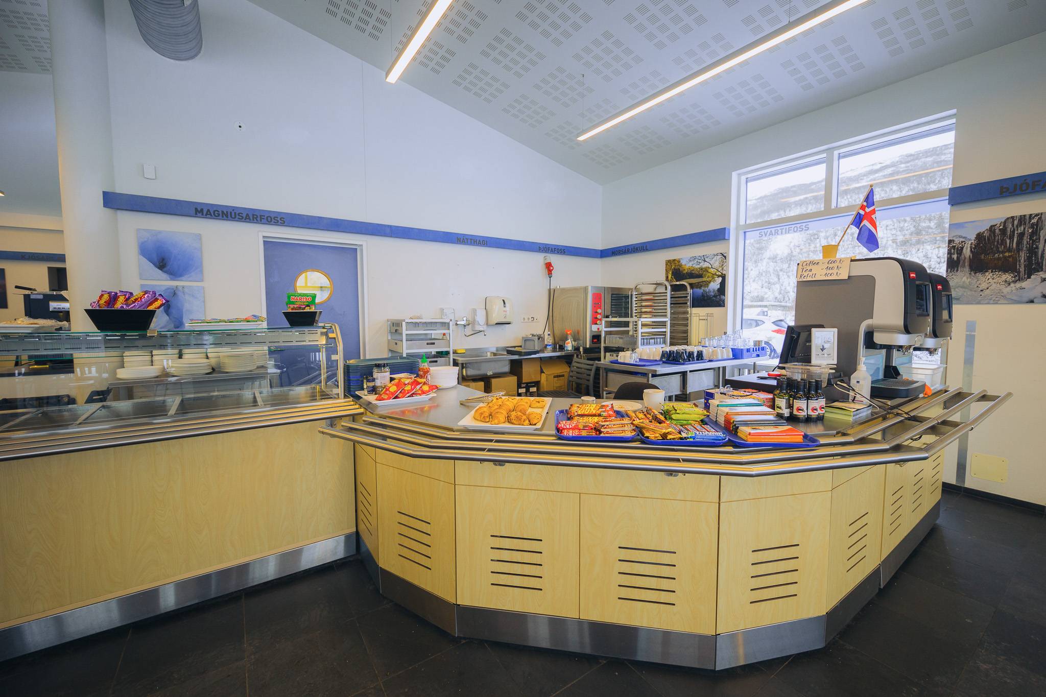 Interior of a bright, modern cafeteria with a curved serving counter. Assorted pastries and sweets are displayed.