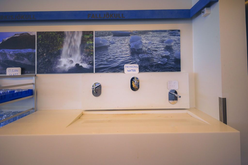 Water station with glacier photos above. Three taps labeled "Drinking Water" and "Out of Order." Clean glasses on the left.