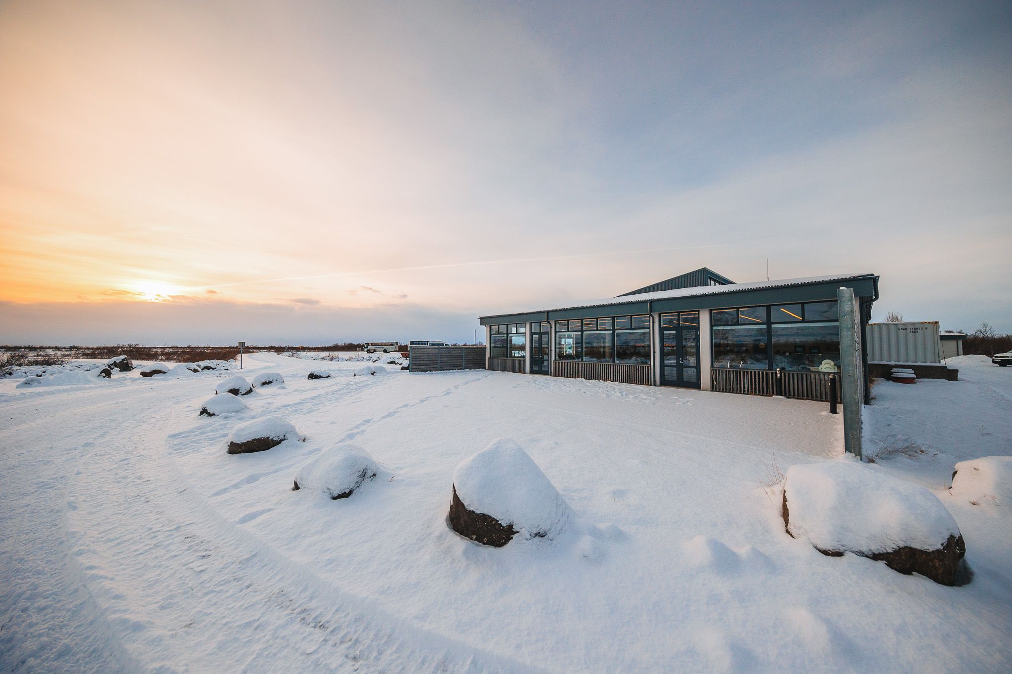 A modern glass building stands in a snowy landscape at sunset, surrounded by snow-covered rocks.