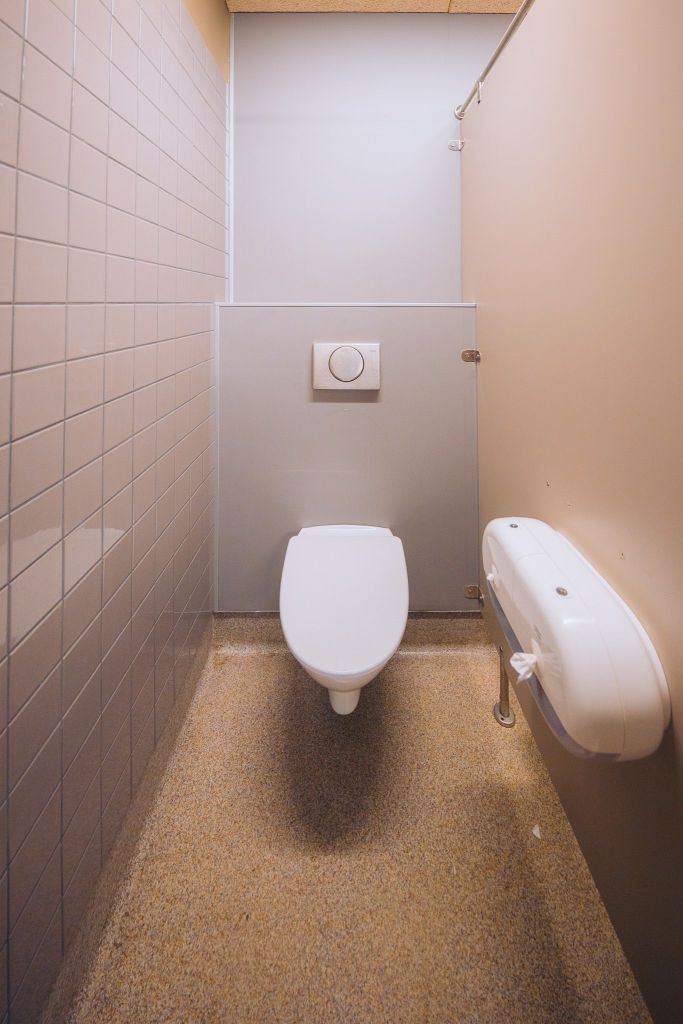 Narrow bathroom stall with beige walls, featuring a wall-mounted toilet and a toilet paper dispenser on the right.