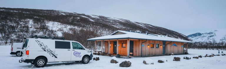Snowy scene with a wooden cabin and a parked white van labeled "CampEasy." Snow-covered hills and mountains form the backdrop under a gray sky.