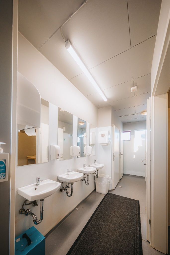 Bright, clean restroom with white sinks and dispensers lined against a wall beneath mirrors. A black mat runs along the gray floor.