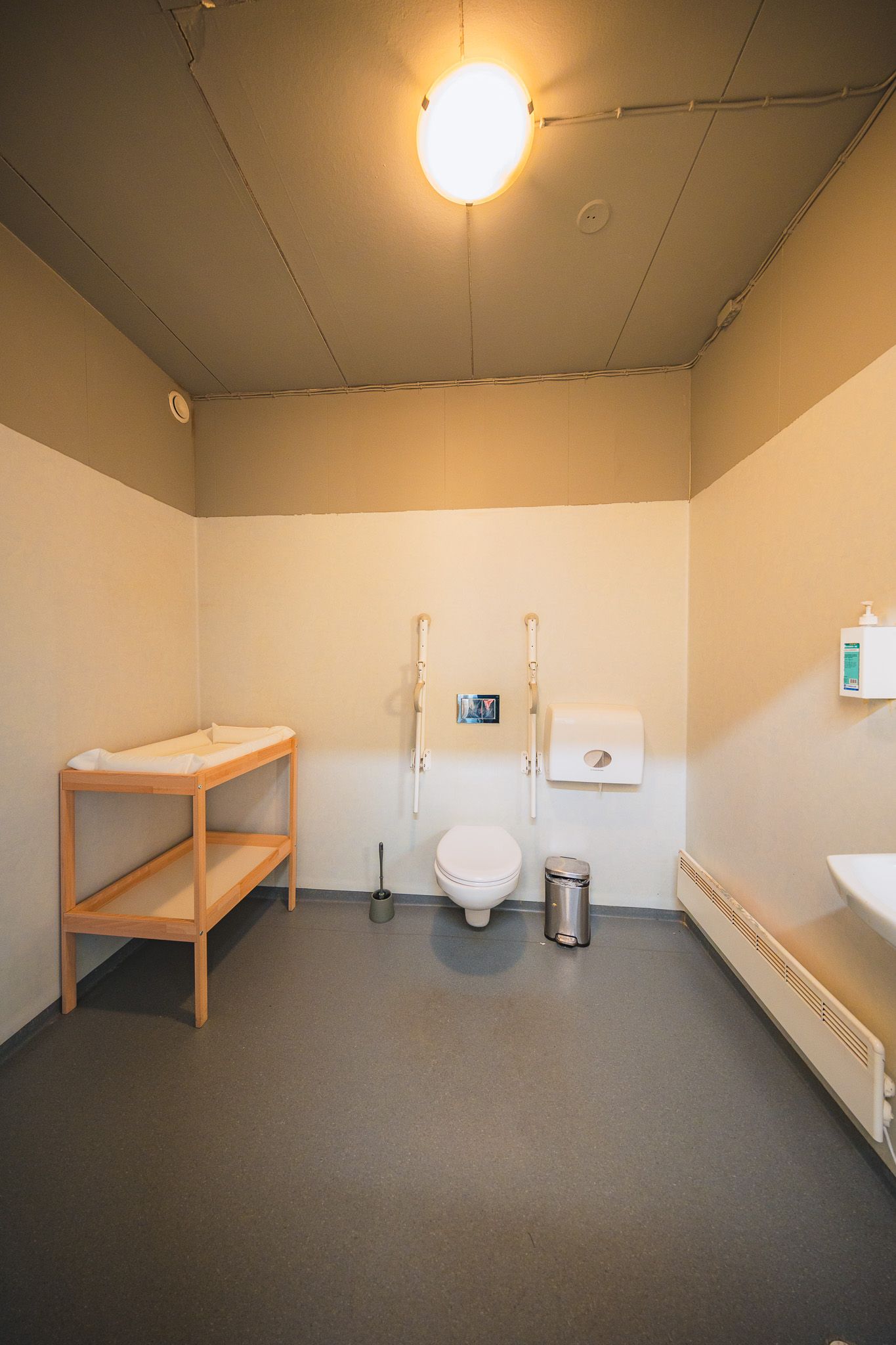 Bright, spacious accessible restroom with grab bars, toilet, changing table on the left, sink on the right, hand sanitizer, and a paper dispenser.
