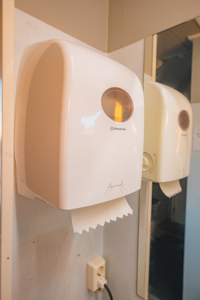 Wall-mounted white paper towel dispenser in a bathroom, with a sheet of paper towel hanging. A mirror reflects the dispenser.