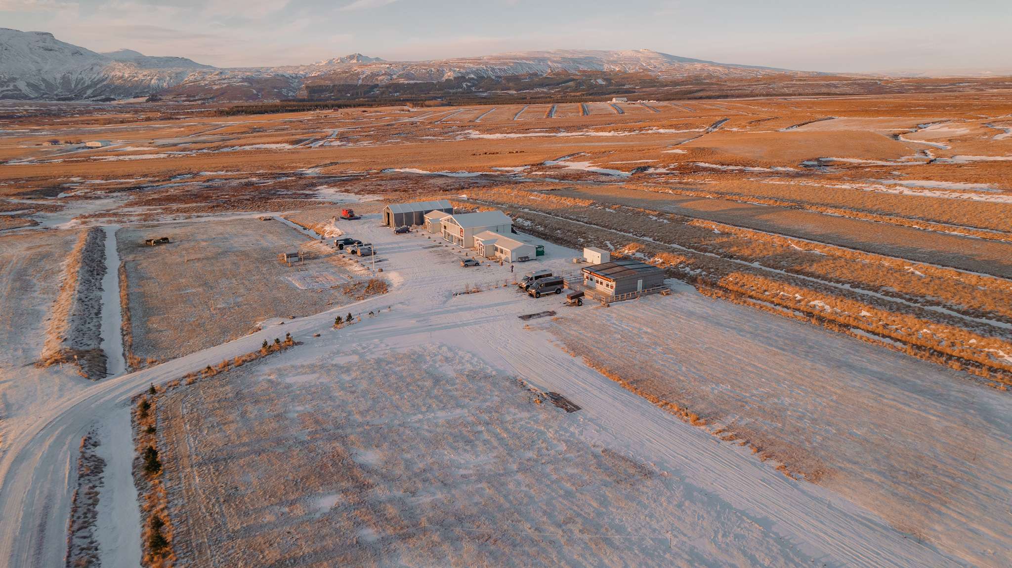 Aerial view of a snowy, rural landscape at sunset. Central farm buildings surrounded by snow-dusted fields and distant mountains under a pink-tinged sky.