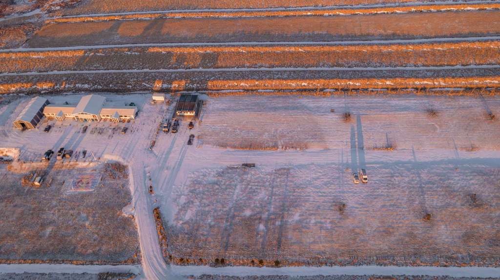Aerial view of snow-covered fields and buildings at sunset. Long shadows stretch across the terrain, casting a warm orange glow over the scene.