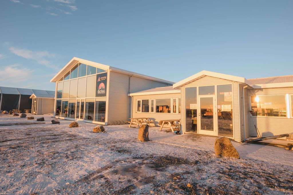 A modern building with large glass windows is bathed in warm sunset light, set against a snowy landscape. Picnic tables and rocks are in the foreground.