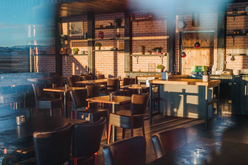 Cozy cafe interior with wooden tables, brown chairs, and warm lighting. Brick walls lined with shelves holding plants and decor, evoking a rustic, inviting vibe.