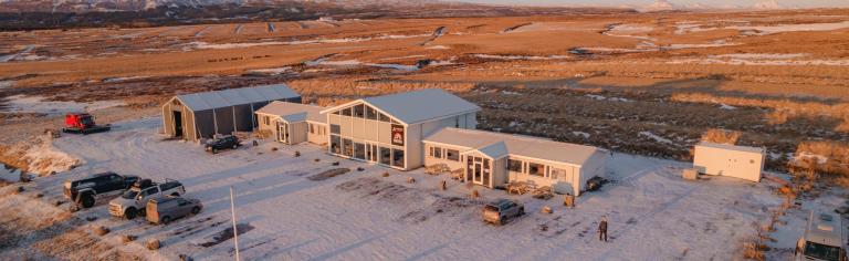 Aerial view of a snow-covered rural building complex at sunrise, casting long shadows. Surrounded by a vast, barren landscape under a clear sky.