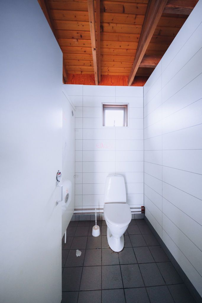 A clean, minimalist bathroom with white tile walls, a white toilet, a toilet brush, and paper.