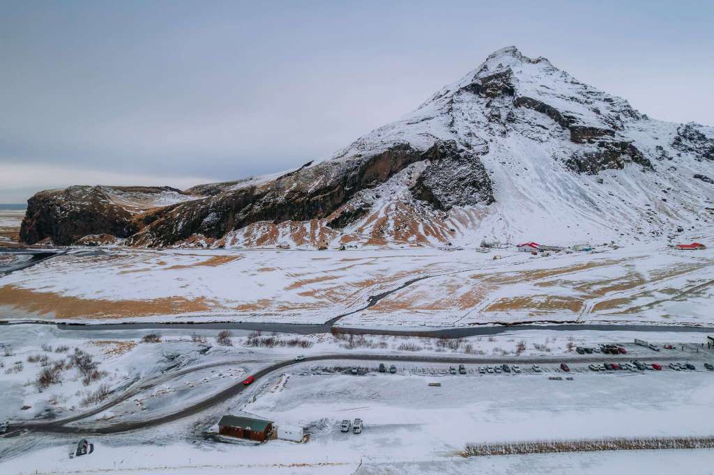 Snow-covered mountain with rugged peaks and a vast, snowy landscape in front. Sparse buildings and a road with vehicles below.
