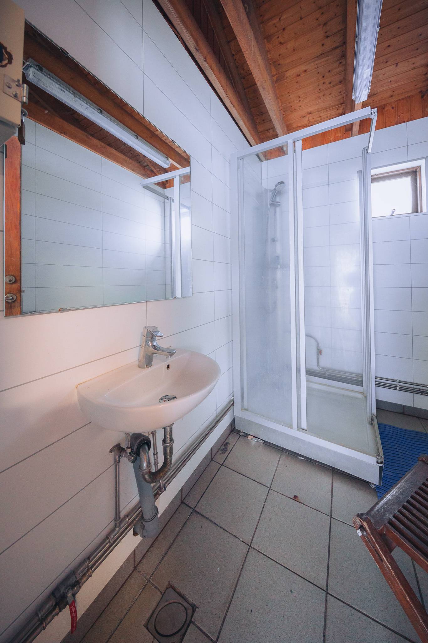 Small bathroom with white tiled walls and gray floor tiles. Features a wall-mounted sink, large mirror, and an enclosed shower.