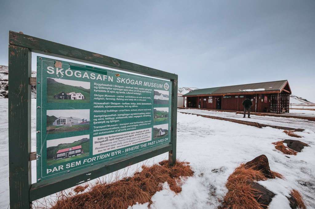 Sign in snowy landscape reads "Skógar Museum," with a rustic building and a lone visitor in the background.