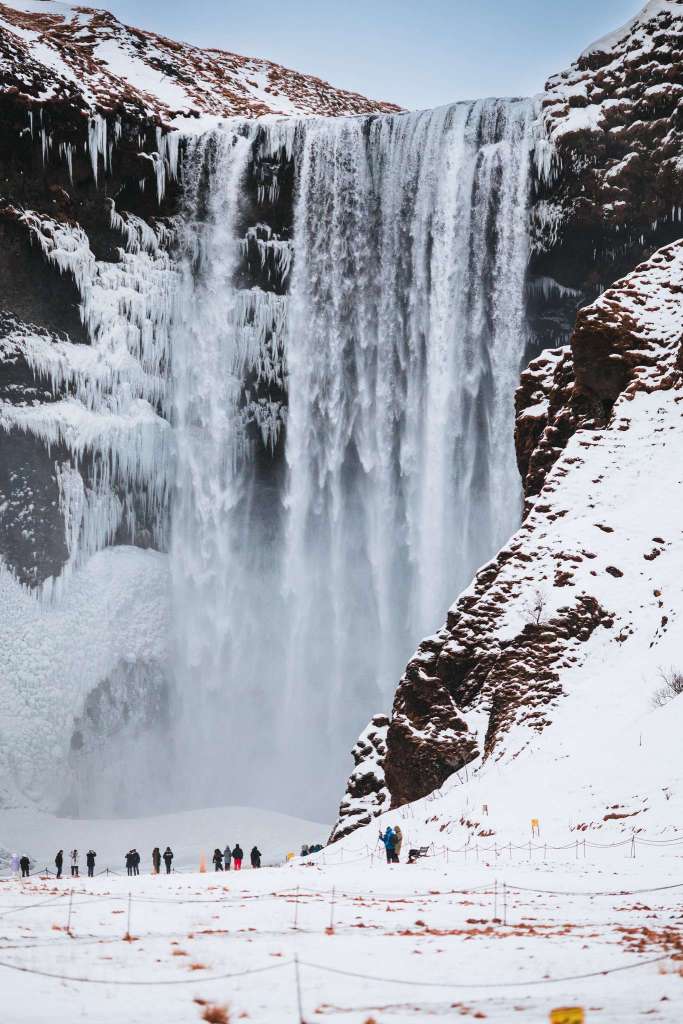 A majestic waterfall cascades down a steep rock face, partially frozen with icicles. People in winter attire stand below on snowy ground.
