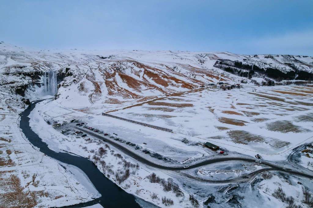 Aerial view of a snow-covered landscape with a winding river and waterfall to the left. Several parked cars line a road, set against snowy hills.