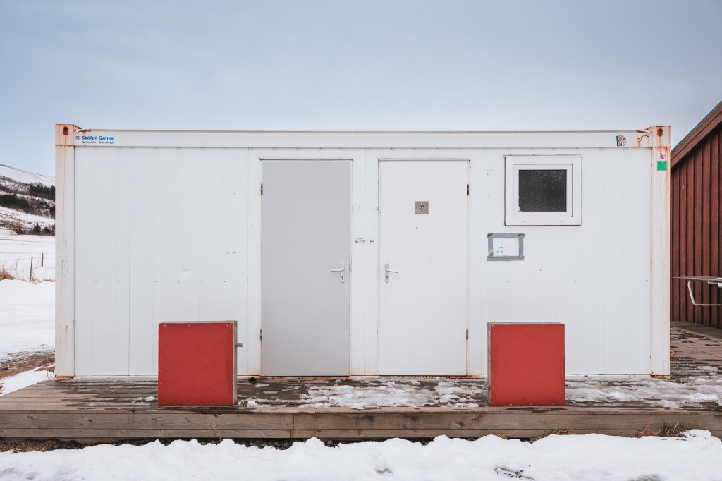 A white container building with two doors and a small window sits on a snowy wooden platform. Two red barriers stand in front, against a gray sky.