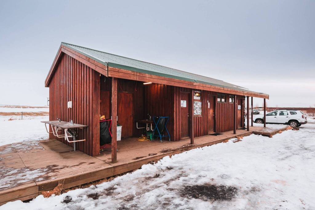 A rustic wooden cabin with a green roof stands on a snow-covered landscape. A white car is parked nearby.