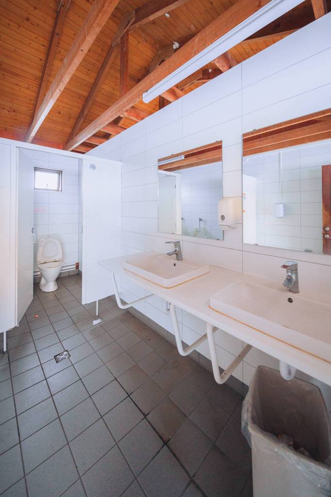 Public restroom with a wooden ceiling and tiled floors. Features two sinks with mirrors, a wall-mounted soap dispenser, and a nearby stall with a toilet.