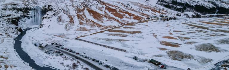 Aerial view of a snowy landscape featuring a waterfall on the left with flowing water, surrounded by snow-covered fields and parked cars in a lot below.