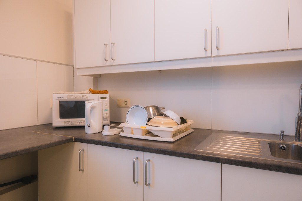 A tidy kitchen corner with white cabinets, a microwave, and a kettle on the counter. Dishes are neatly stacked in a drying rack by the sink.
