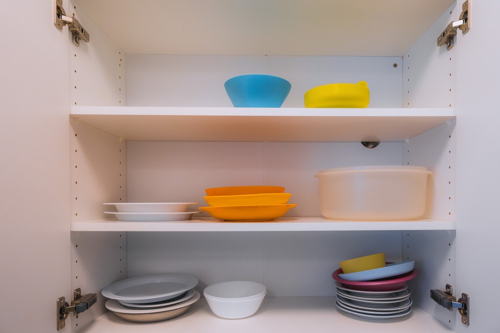 Open white kitchen cabinet with three shelves, displaying various colorful plastic bowls, plates, and a container.