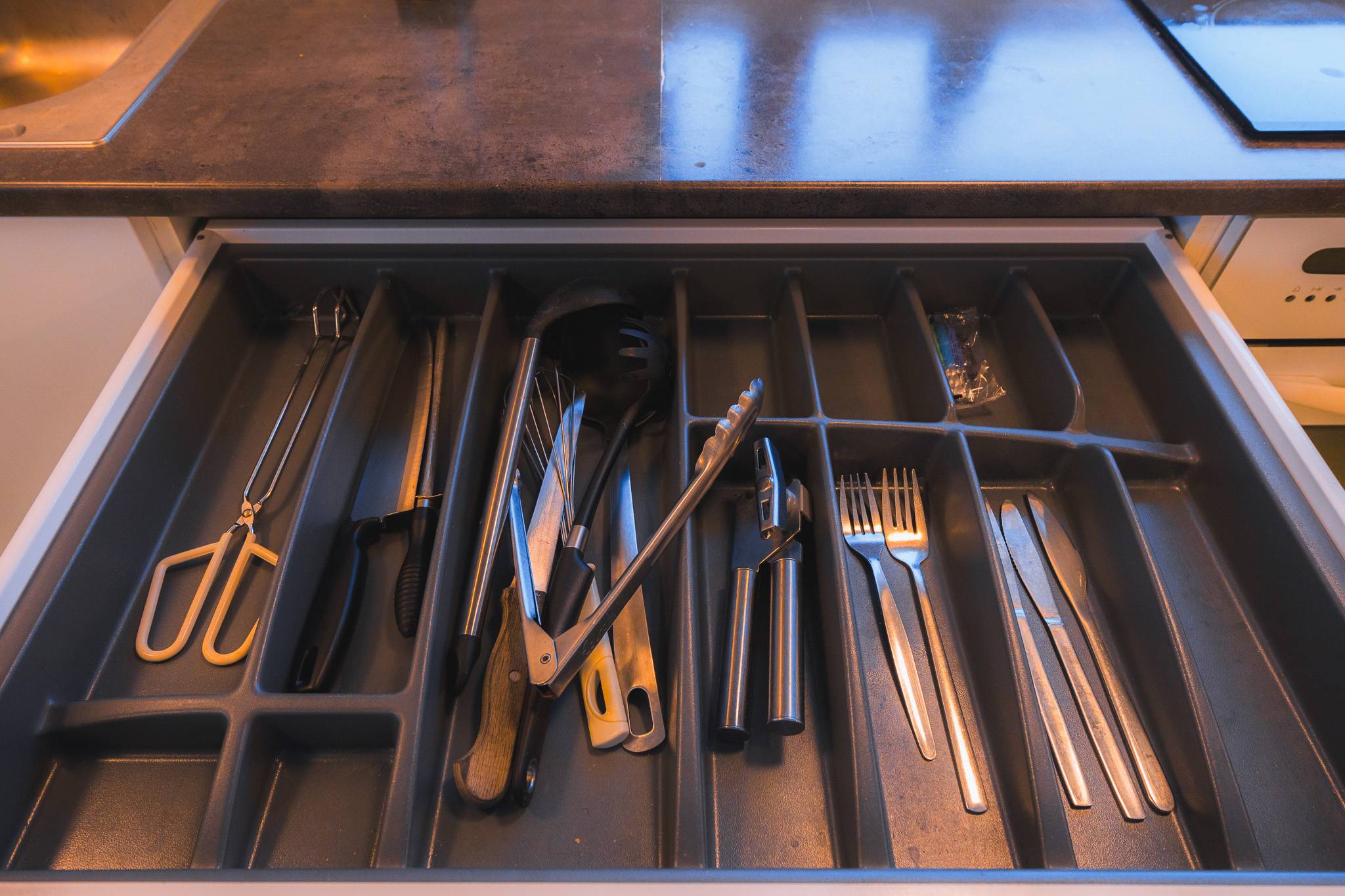 Open kitchen drawer with organized utensils and cutlery in a dark countertop. Contains tongs, scissors, spoons, forks, knives, and other kitchen tools.