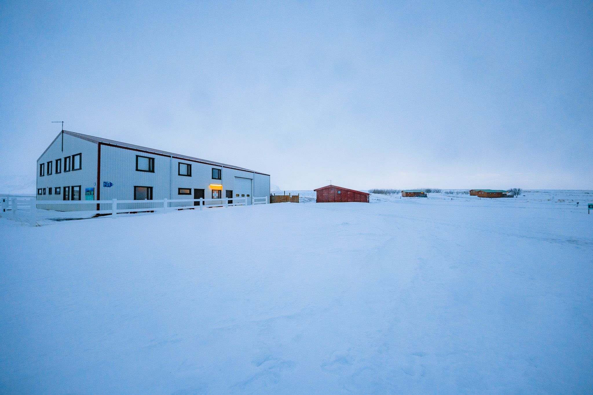 A large white building with dark windows sits against a vast, snow-covered landscape under a cloudy sky. A few smaller red barns are in the distance.