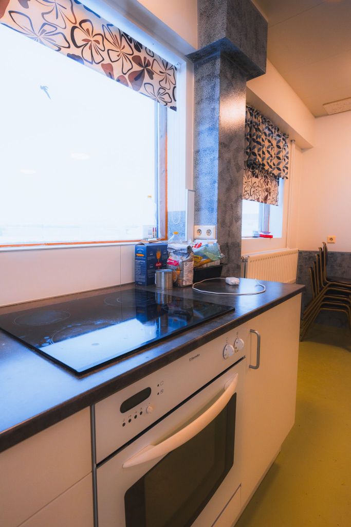 Bright kitchen with a black electric stove and oven. Shelves hold a cereal box and canned goods. Window with floral curtains adds warmth.