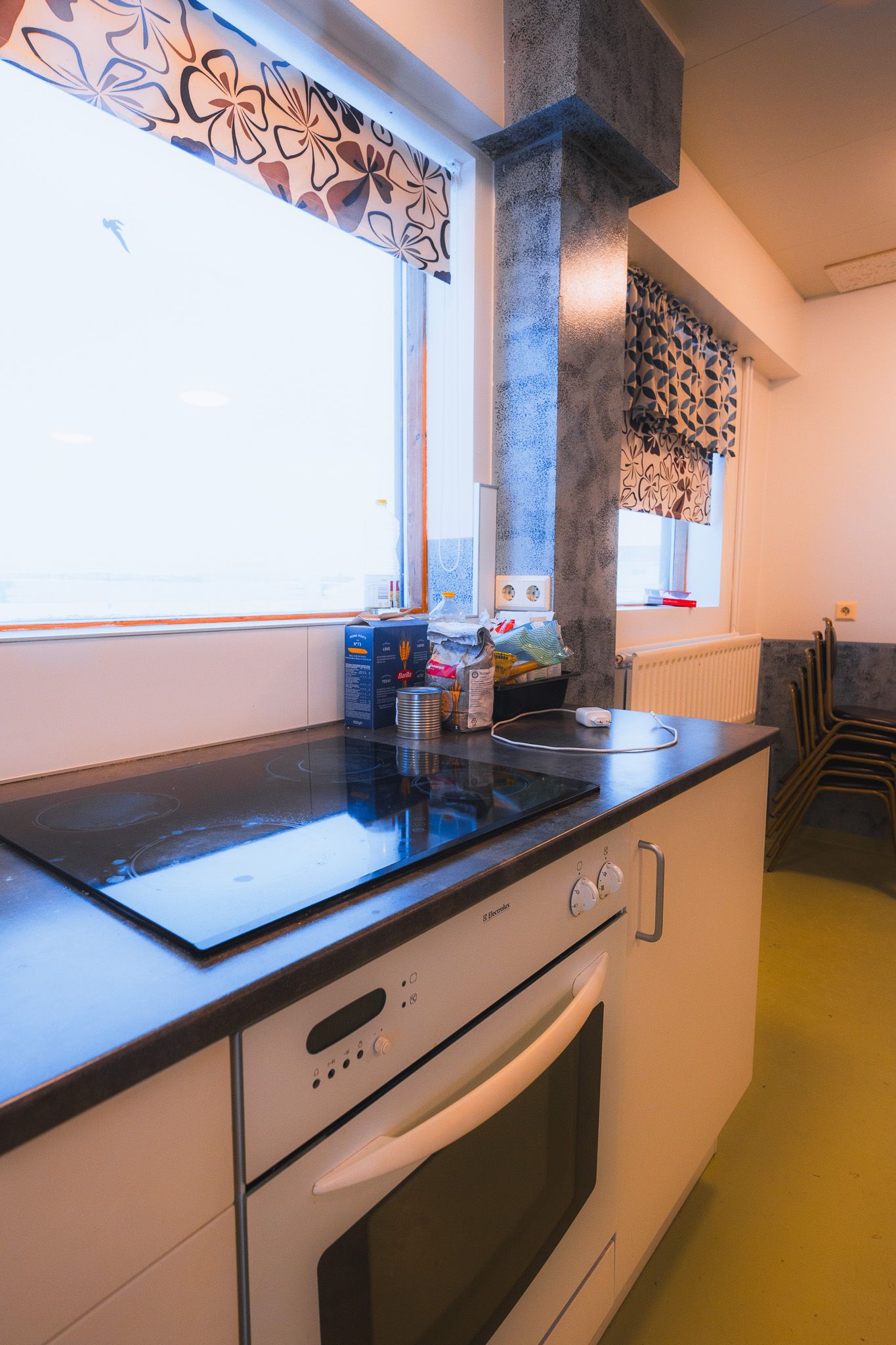 Bright kitchen with a black electric stove and oven. Shelves hold a cereal box and canned goods. Window with floral curtains adds warmth.