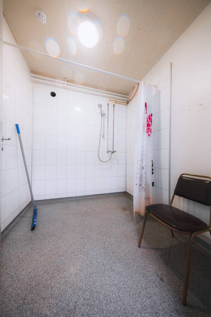 Minimalist bathroom featuring white tiled walls, a showerhead, and a colorful curtain. A broom and chair rest on the gray floor.
