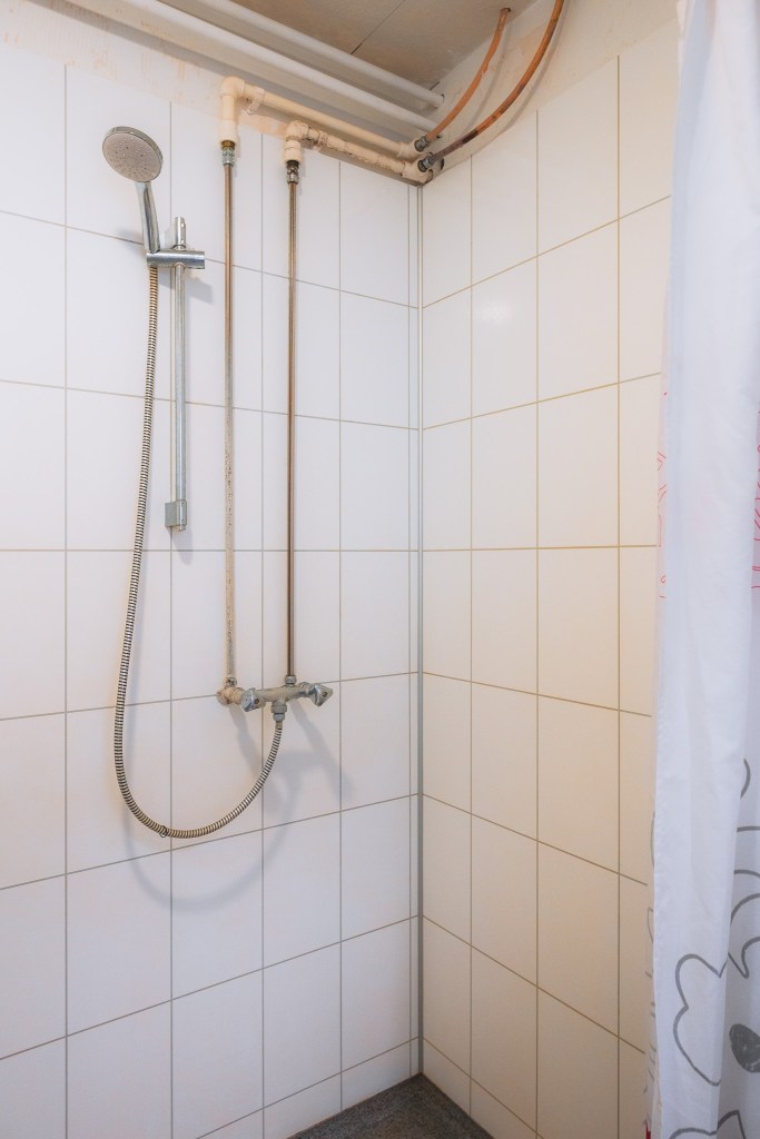 Plain tiled shower with a handheld showerhead attached to the wall. Exposed pipes run along the ceiling and down the wall. A curtain is partially visible.