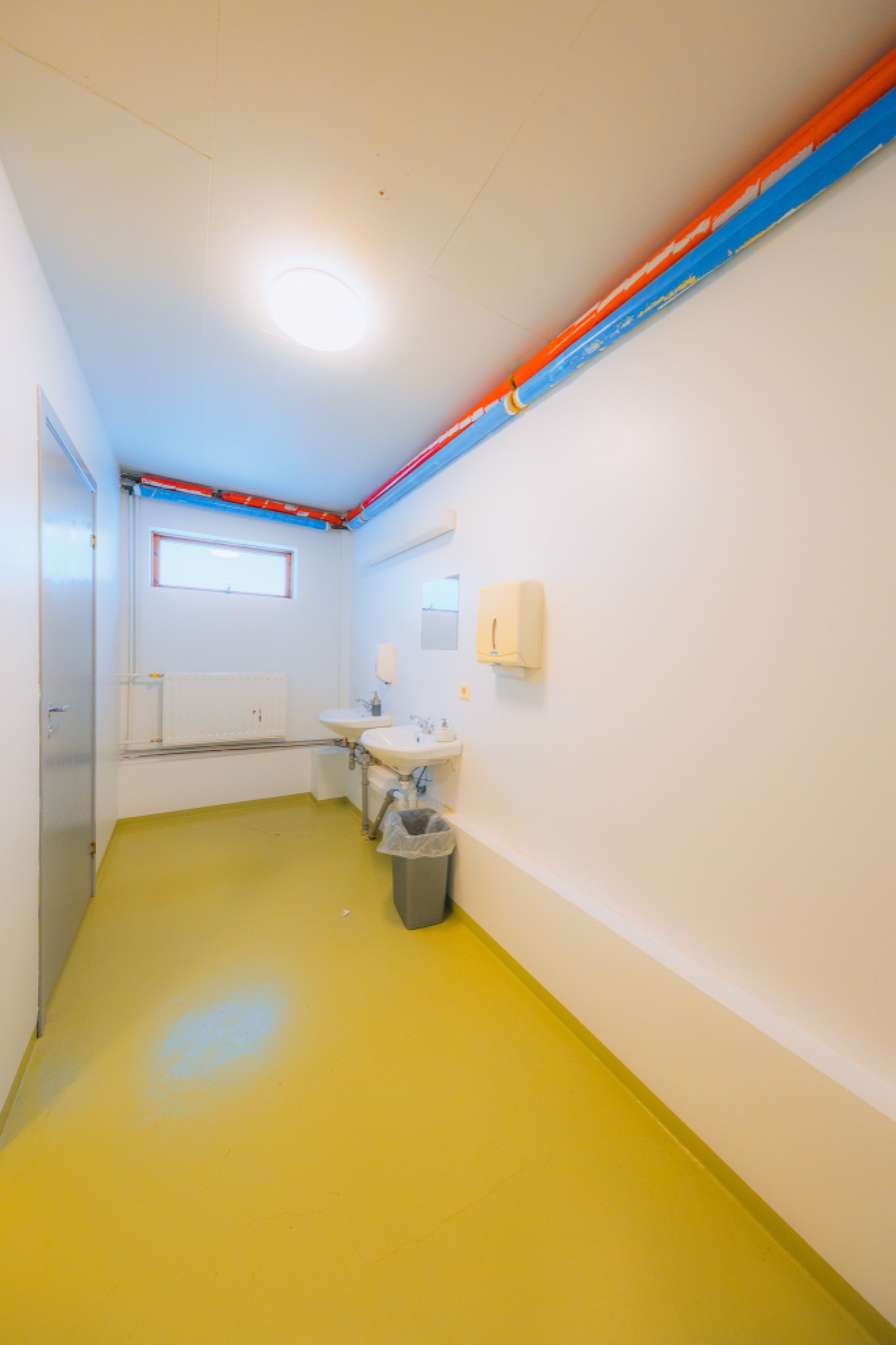 Narrow, brightly lit room with yellow floor and white walls. Colorful pipes run along the ceiling. Features a sink, mirror, and trash bin.