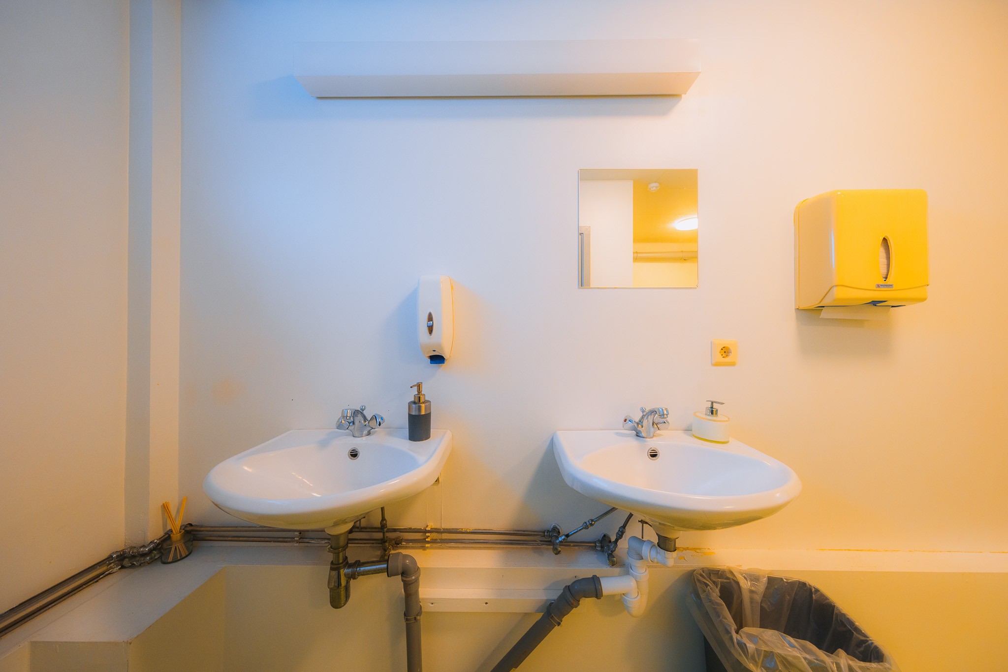Two white sinks with silver faucets are mounted on a pale yellow wall. A soap dispenser and a yellow paper towel holder are above them, while a trash bin is below.