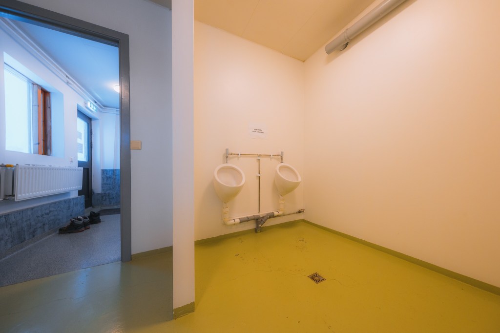 Two urinals are mounted on an empty beige wall. To the left, an open door reveals a dimly lit corridor with a radiator and shoes on the floor.