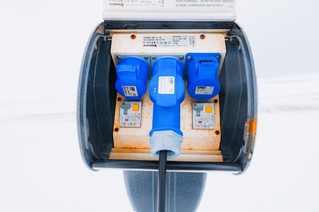 Close-up of a blue electrical connector plugged into a weathered outlet box with three sockets, set against a snowy background.