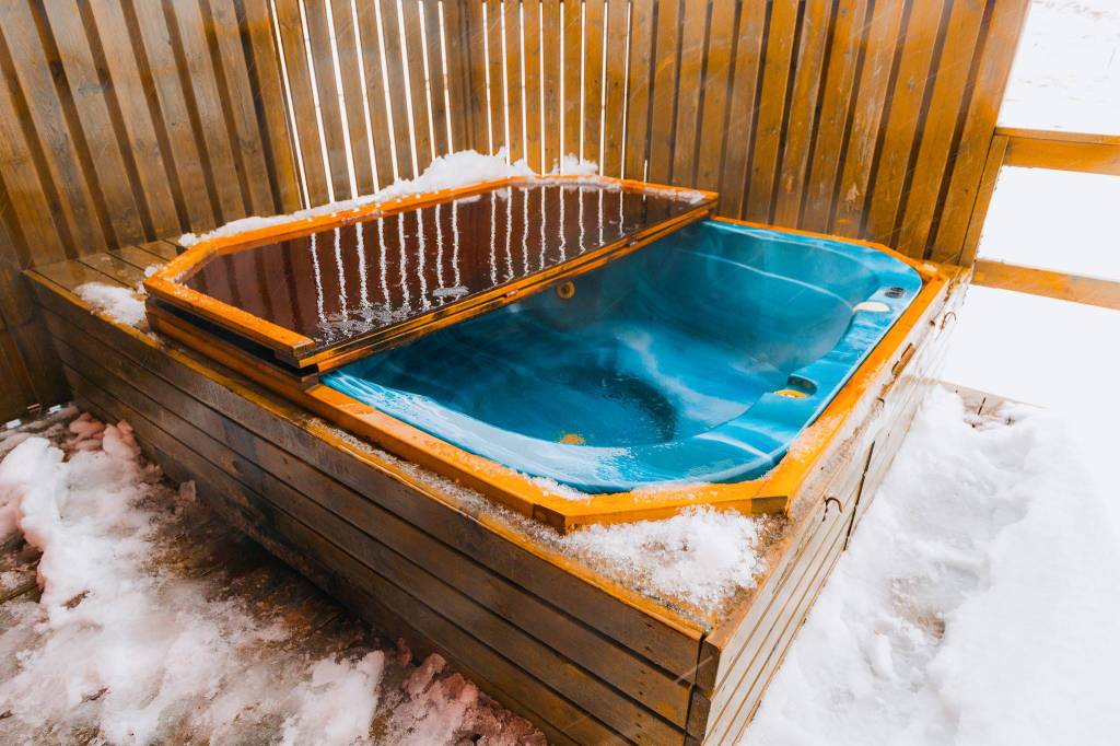 Wooden outdoor hot tub with a partially open cover, surrounded by snow.