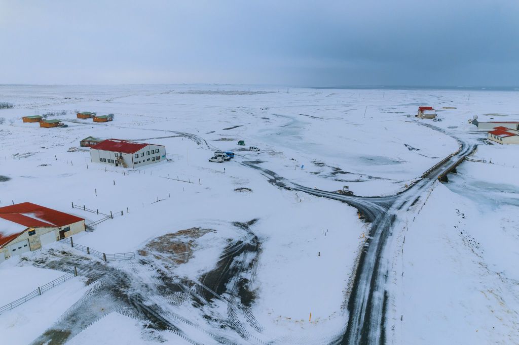 A snowy landscape with scattered red-roofed buildings and winding roads creates a serene, isolated atmosphere under a vast, gray sky.