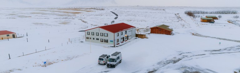 Snow-covered landscape with a central building featuring a red roof. Two parked vans in front. Surrounding area has scattered buildings and barren fields.