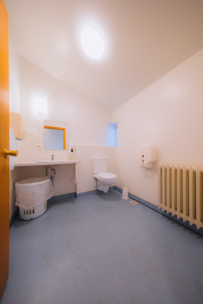 A clean, minimalist bathroom with white walls and a blue floor. Features include a toilet, sink, mirror, small window, radiator, and waste bin.