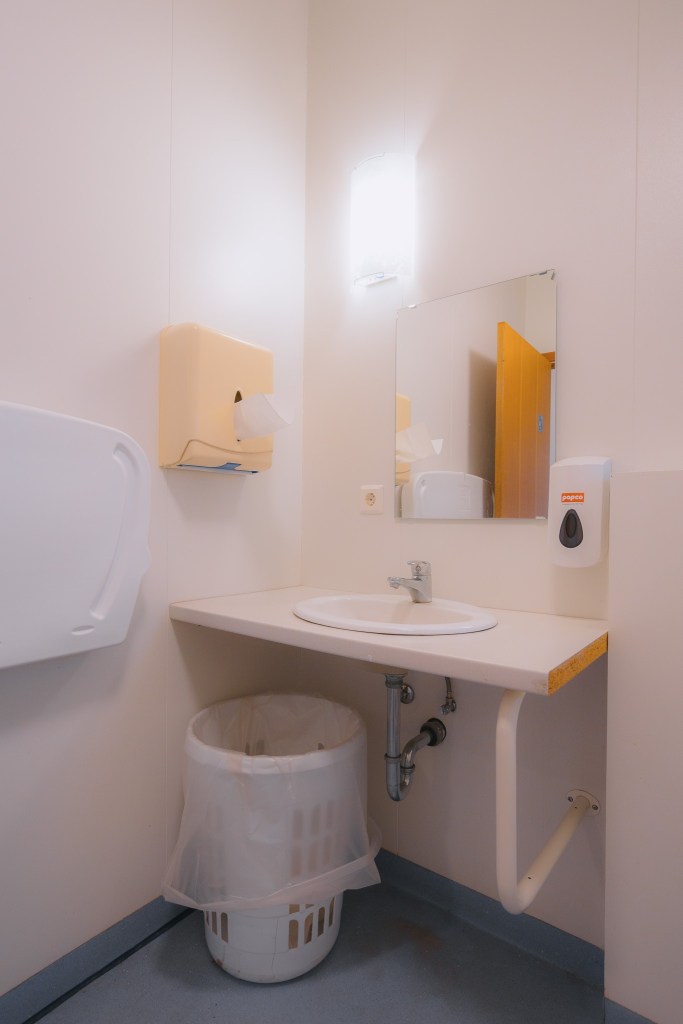 A bright, clean bathroom with white walls features a sink, mirror, soap dispenser, paper towel holder, and a wastebasket beneath the counter.
