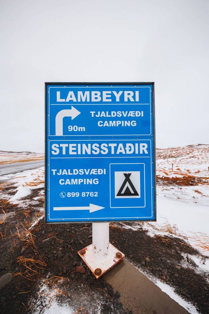 Blue camping sign with snow-dusted terrain in the background. It directs to "Lambeyri" and "Steinsstaðir".