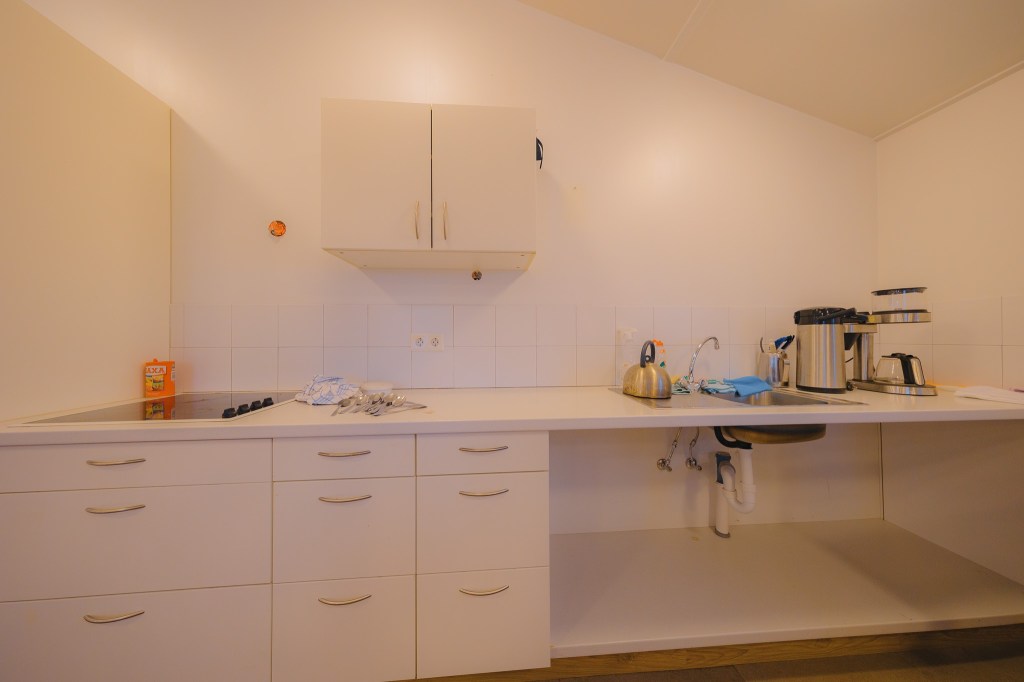 A clean, minimalist kitchen features white cabinets, a stovetop, a sink with a kettle, and a coffee maker.