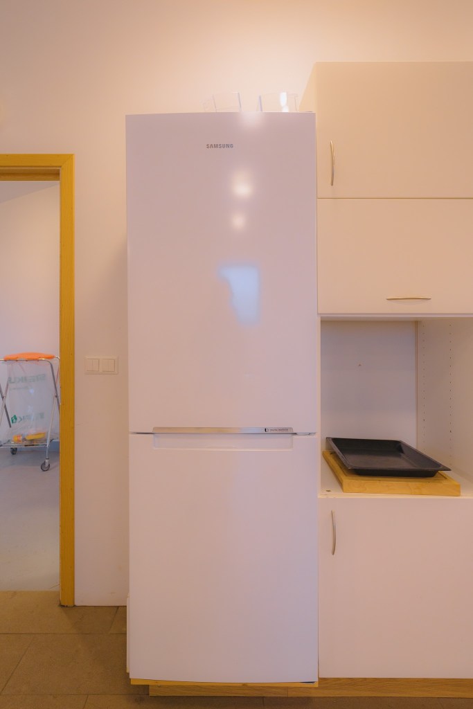 A white refrigerator stands between a kitchen cabinet and a doorway. Soft indoor lighting casts a warm tone. A large tray is on a counter beside the fridge.